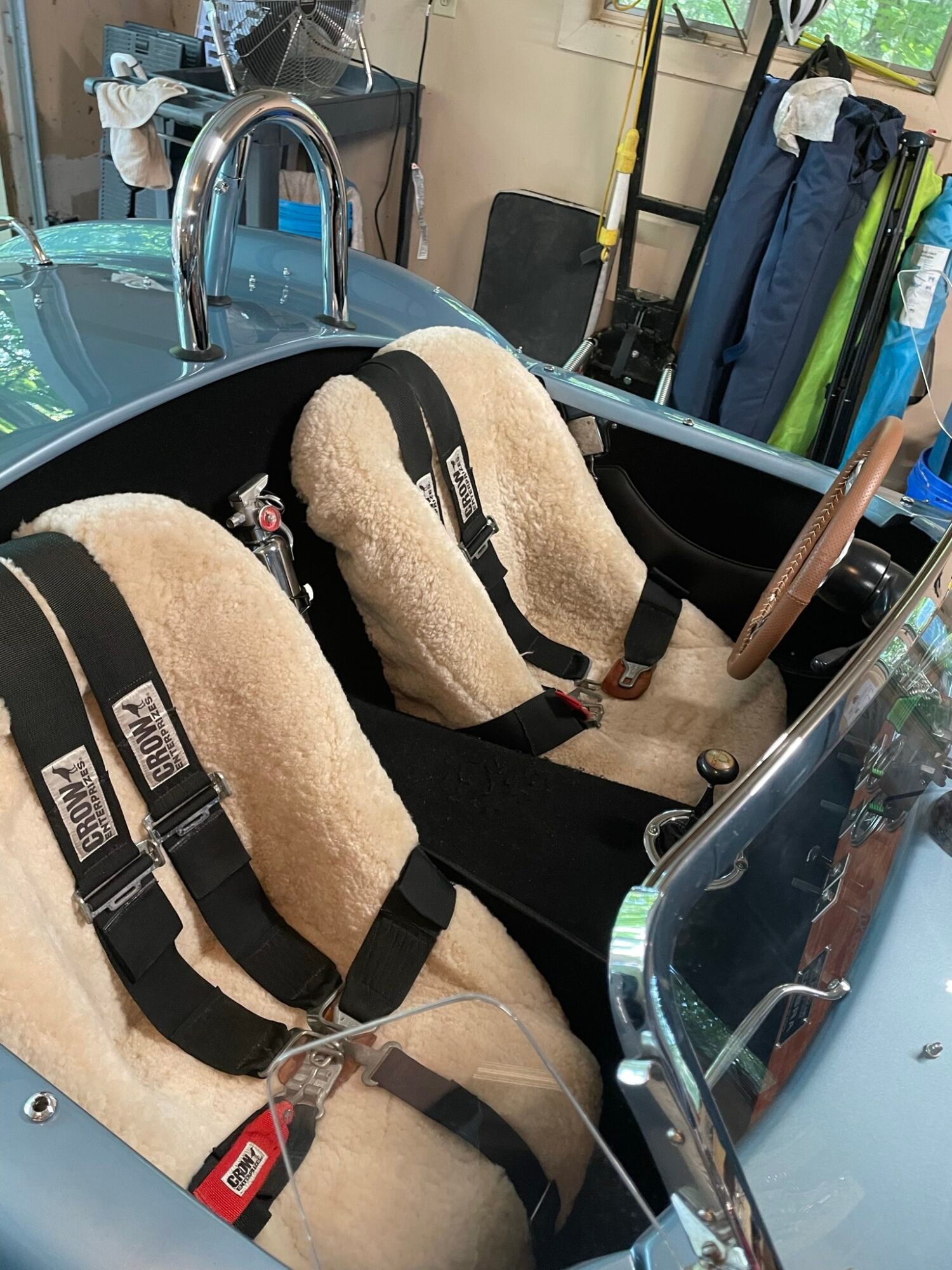 interior of a classic sports car, possibly a Shelby Cobra replica. The seats are covered in sheepskin seat covers and are equipped with racing harnesses.