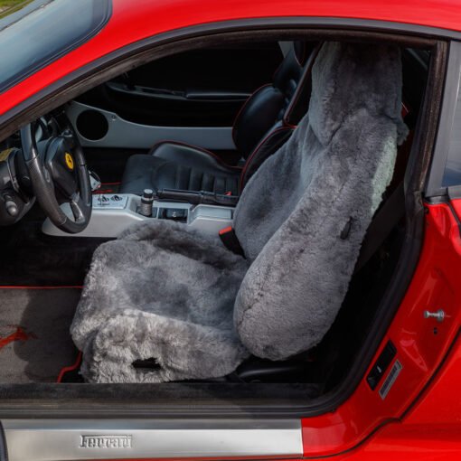 The image shows the interior of a red Ferrari sports car. The driver's seat is covered with a gray sheepskin seat cover. The car has a black leather interior with red stitching.